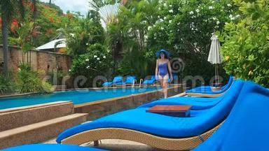 在阳光明媚的日子里，穿着蓝色泳衣、帽子和太阳镜的漂亮女士带着清澈的水在游泳池里走来走去。 <strong>奢华</strong>